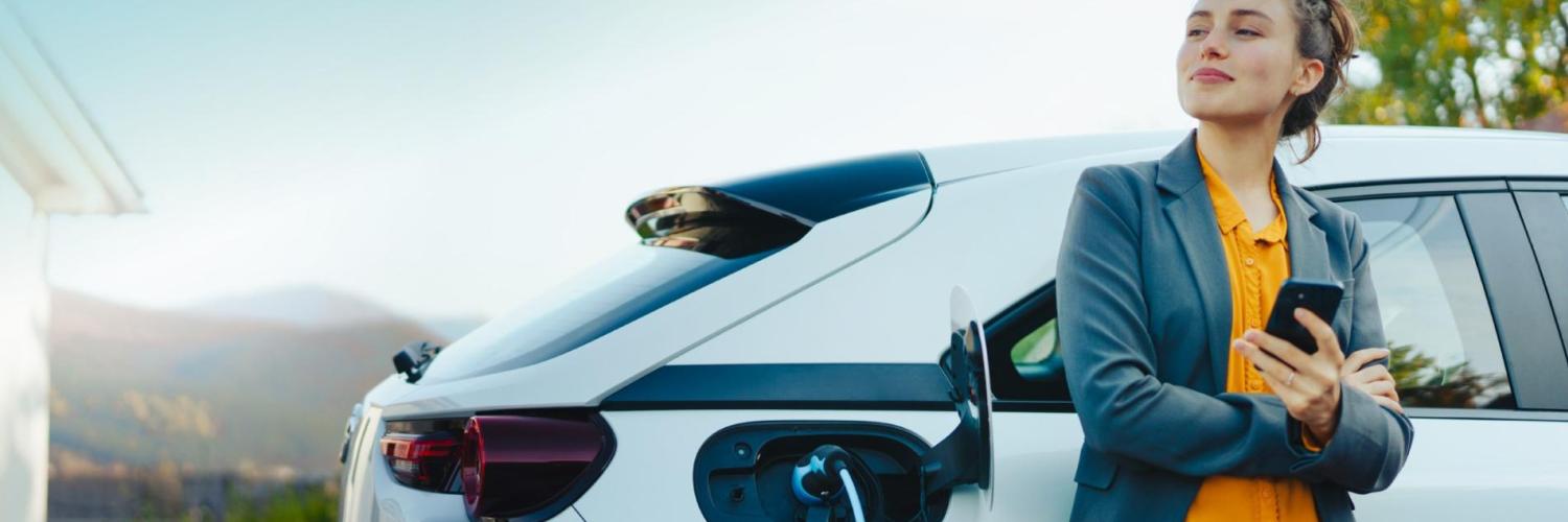 Arval Flex-Frau vor ihrem Elektroauto Frau in gelbem Kleid lehnt an ihrem Elektroauto während des Ladevorgangs und genießt die flexible Langzeitmiete von Arval